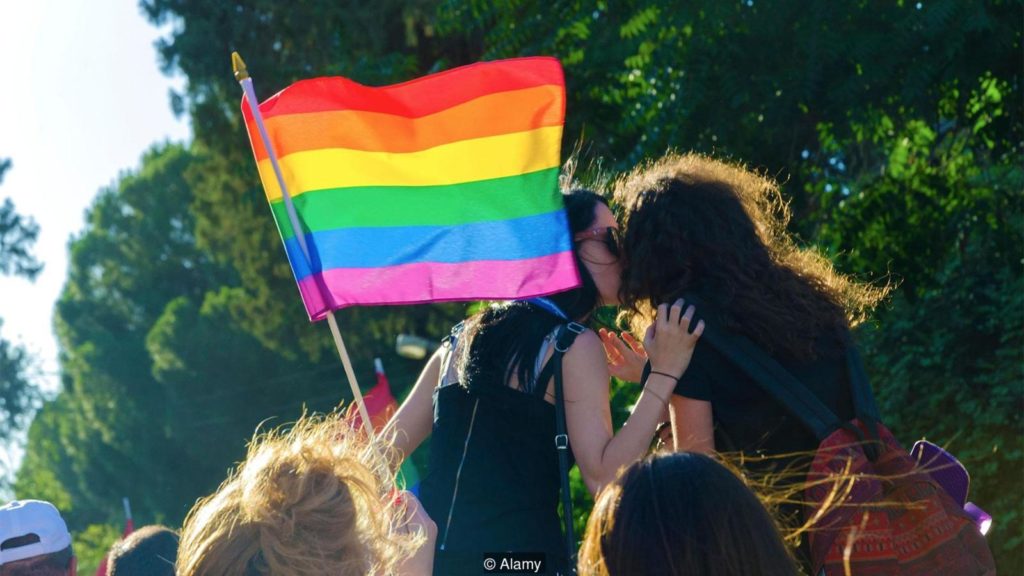 Duas garotas se beijando ao lado de uma bandeira do arco-íris.